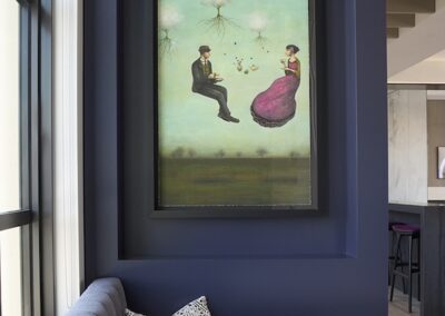 A framed surreal painting of a man and woman floating with trees above them hangs on a dark blue wall above a gray cushioned bench and a patterned pillow near a window.