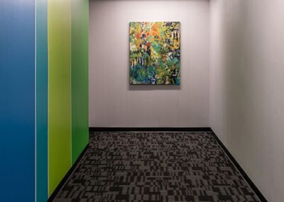 A hallway with patterned carpet, light grey walls, blue and green panels on the left, and a colorful abstract painting hanging at the end wall.