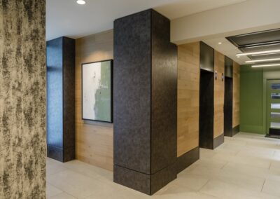 Modern building lobby with wood-paneled walls, two black elevator doors, abstract wall art, tiled floor, and green accent wall near the entrance.
