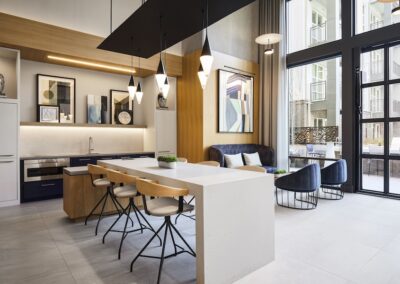 Modern kitchen and lounge area with a white island, wooden chairs, pendant lights, wall art, large windows, and outdoor patio visible.
