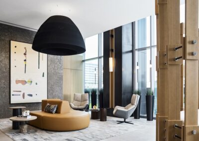 Modern lobby with tan circular seating, abstract wall art, tall windows, black pendant lamp, armchairs, and indoor plants.
