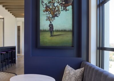 A framed surreal painting of a man with floating objects above his head hangs on a dark blue wall in a modern seating area with a table, bench, and window.