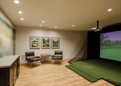 A room with a golf simulator, seating area with two chairs and a table, framed golf course art on the wall, and wood flooring.