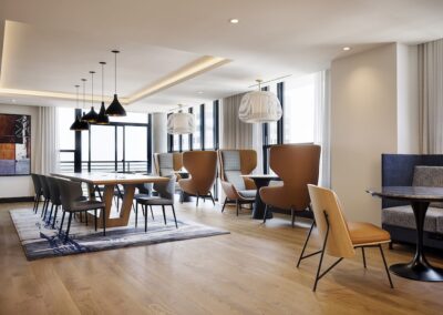 Modern open-concept lounge with long table, mixed seating including high-back chairs, pendant lighting, large windows, and hardwood floors.