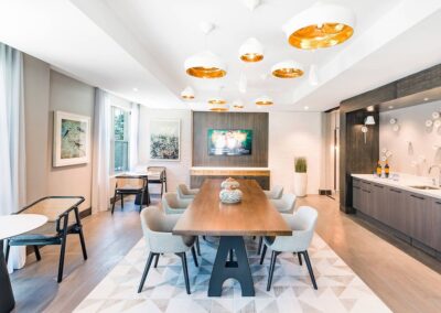 Modern conference room with a long wooden table, eight chairs, pendant lights, wall art, a TV, and a kitchenette area with drinks on the counter.