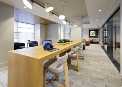 A modern office lounge with a long wooden table, several chairs, a laptop on the table, and a seating area in the background.
