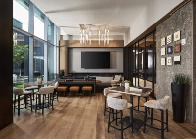 Modern lounge area with wooden flooring, tables and chairs, wall-mounted TV, decorative wall art, and floor-to-ceiling windows letting in natural light.