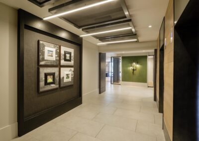 A modern hallway with framed pictures on a dark accent wall, beige floor tiles, and a view into another room with green walls and a wall sconce.