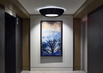 A hallway with two elevators, neutral tile flooring, dark wall panels, and a backlit painting of a tree on the wall under a large circular ceiling light.