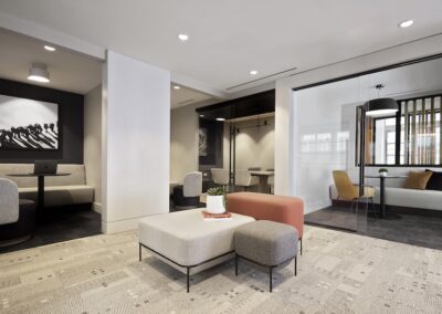 Modern office lounge with modular seating, a small table with a plant, built-in booths, and a glass-walled meeting room with armchairs. Neutral tones and minimalist decor throughout.