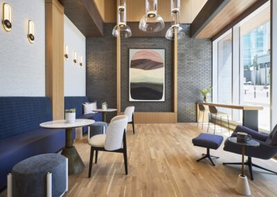 Modern lounge area with wooden floors, blue seating, small tables, abstract wall art, and large floor-to-ceiling windows letting in natural light.