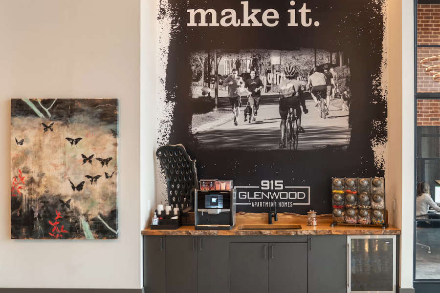 A coffee station with a machine, condiments, and a mini fridge sits below a large wall mural of cyclists and a painting with butterflies on a nearby wall.