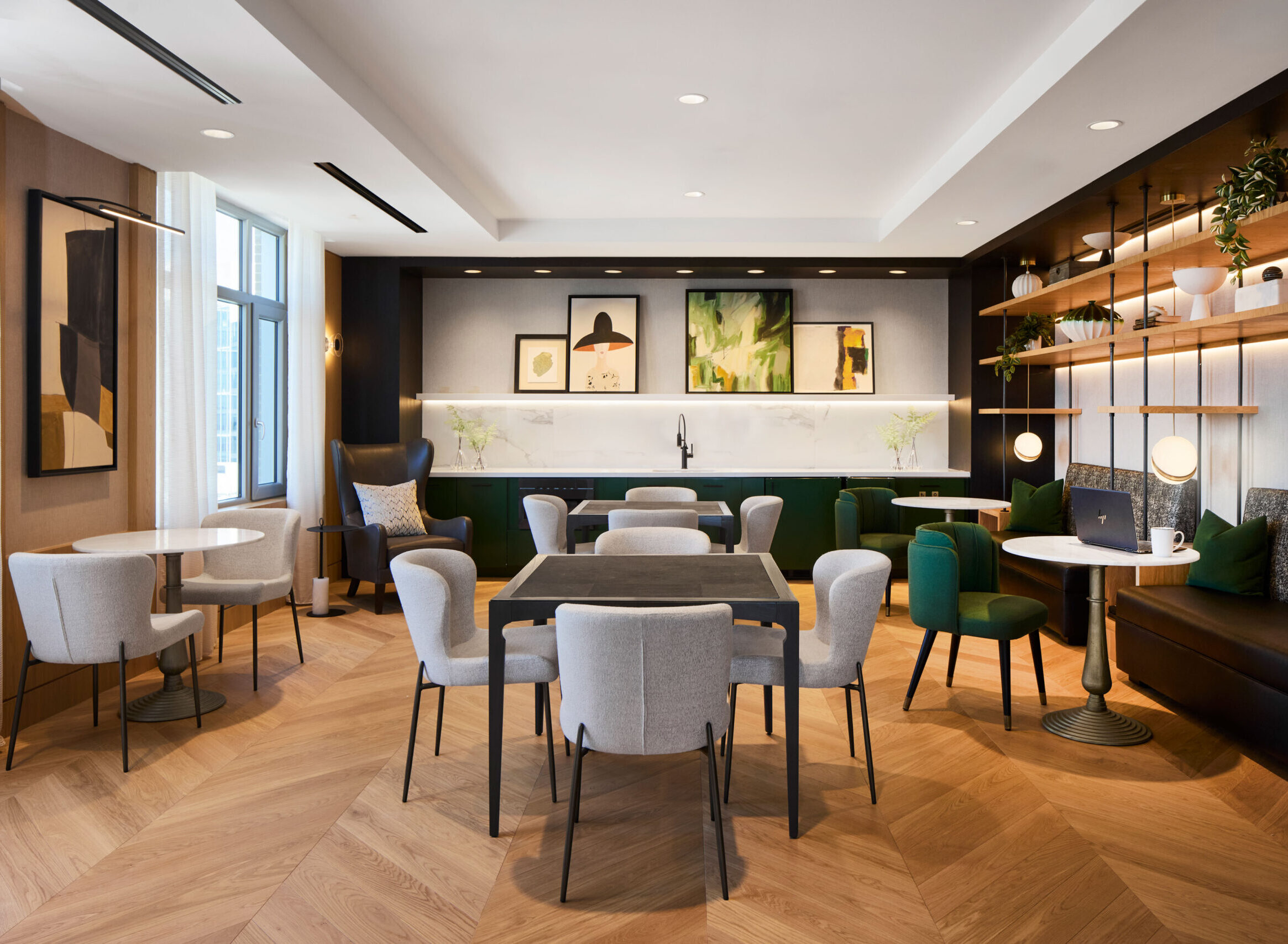 Modern lounge area with tables, chairs, wall art, plants, and a built-in bench under warm lighting; wood herringbone floor and large windows provide a bright, inviting atmosphere.